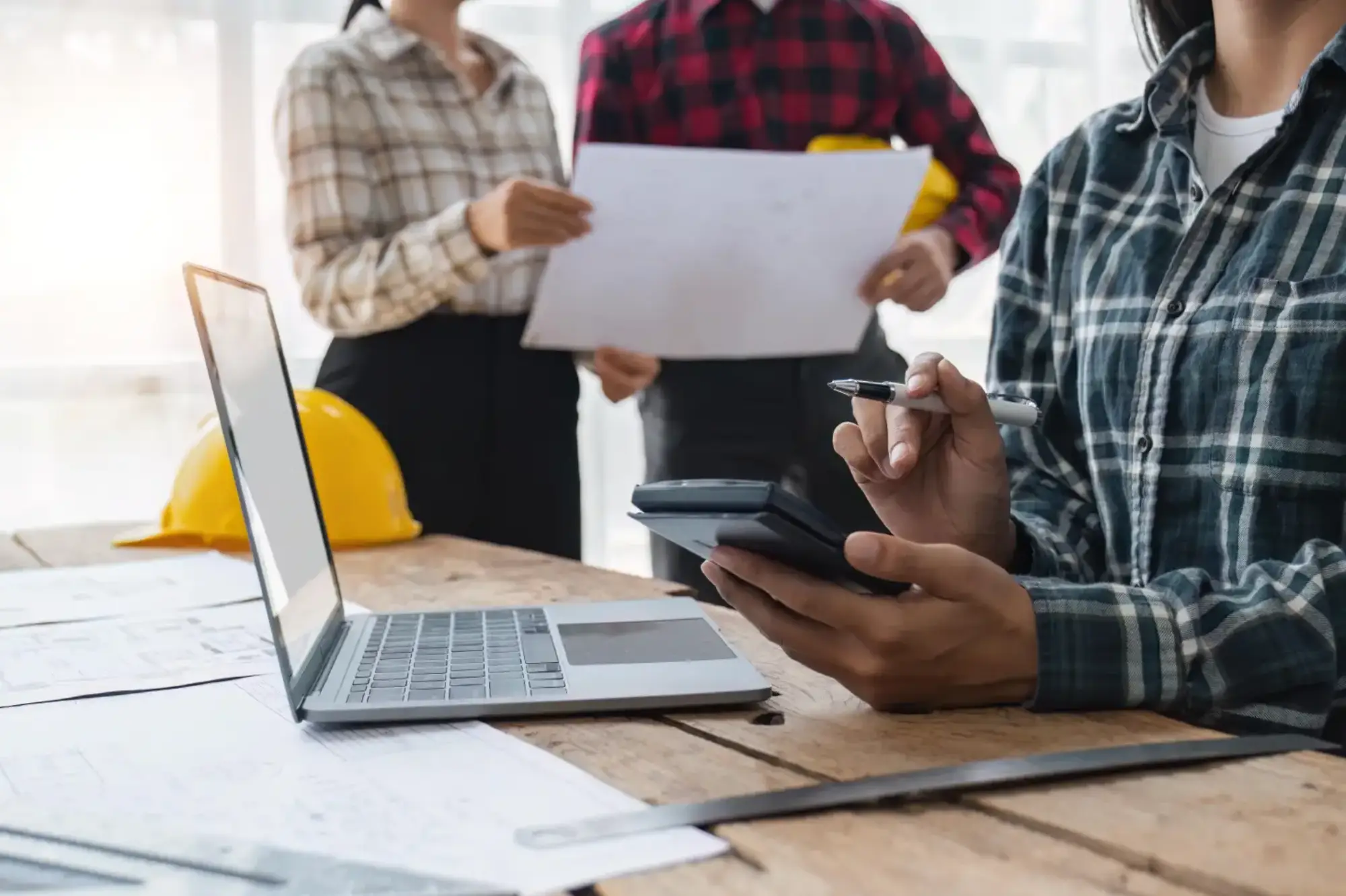 Trois professionnels en chemises à carreaux examinent des plans de construction à une table avec un ordinateur portable, une calculatrice et des casques de sécurité.