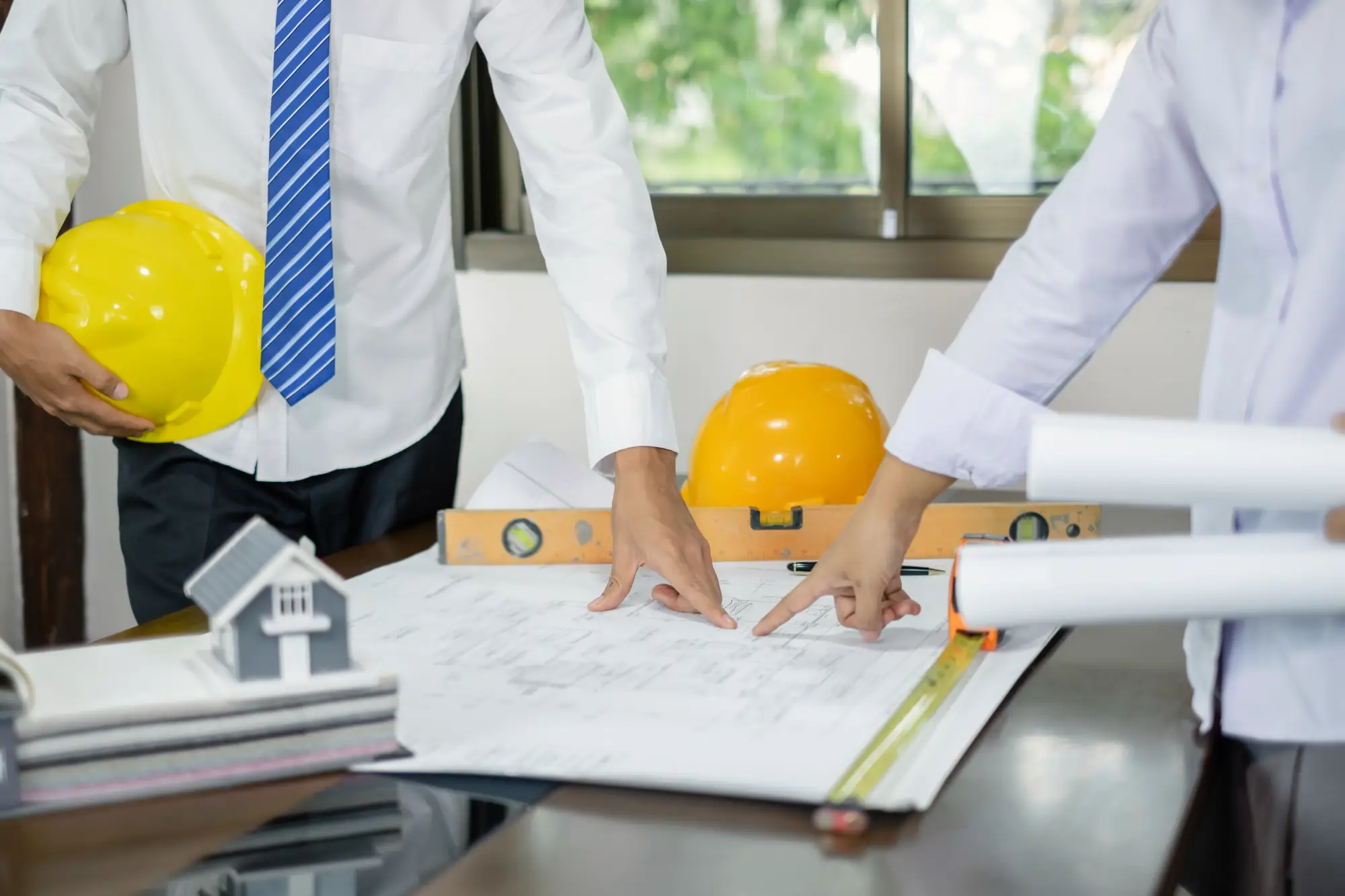 Deux professionnels de la construction analysent des plans sur une table, discutant des détails tout en tenant des outils de construction et des casques de sécurité.