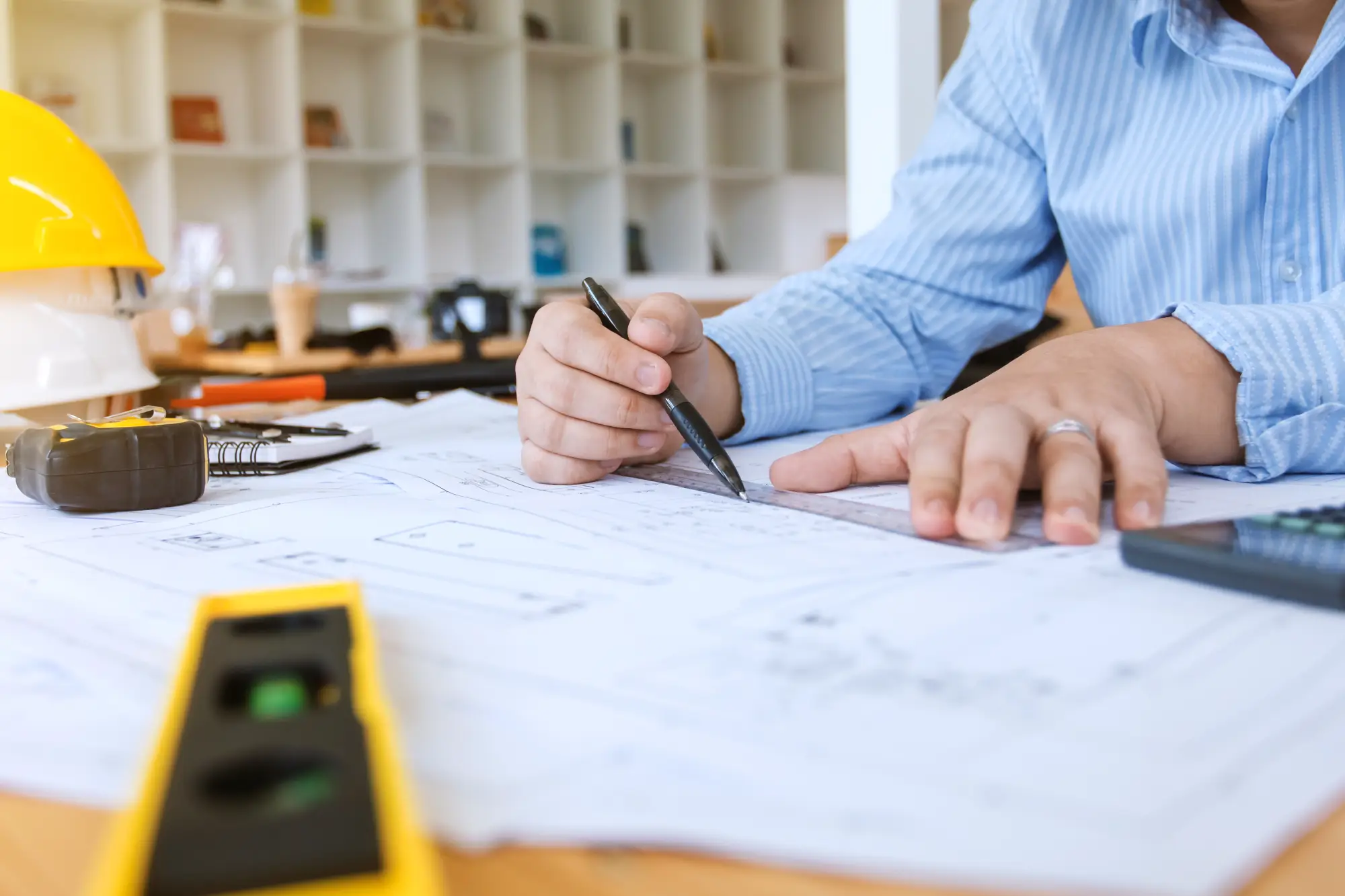 Une personne en chemise rayée examine des plans architecturaux avec une règle, entourée d'outils dont une calculatrice et un ruban à mesurer.